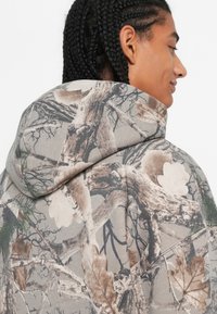 Cilvēks ar pīts matiem, kurš valkā pelēku kapuces džemperi ar detalizētu brūnu un bežīgu koka zara un lapu maskēšanās rakstu.