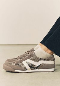 Braune Sneakers mit Schlangenledermuster, weißen Akzenten, aus Wildleder und mit strukturierten Sohlen. Getragen mit hellbeigen Socken und schwarzen Hosen.
