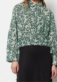 Kobieta w zielonej koszuli z guzikami i ciemnym abstrakcyjnym wzorem w kropki, włożonej w czarną spódnicę, stojąca na jednolitym tle.