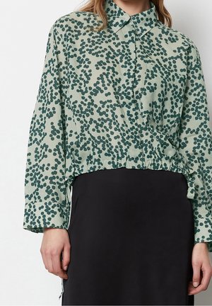 Femme portant une chemise verte à boutons avec un motif abstrait de points sombres, rentrée dans une jupe noire, se tenant devant un fond uni.