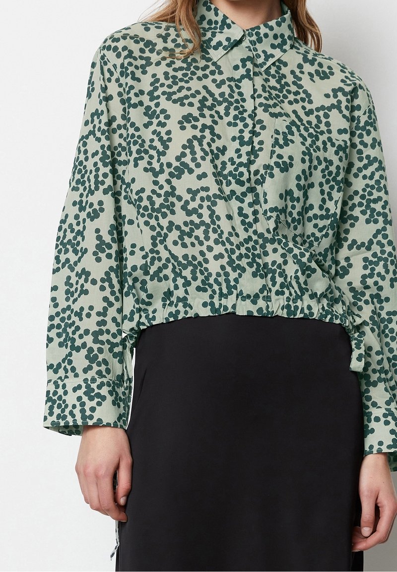 Kobieta w zielonej koszuli z guzikami i ciemnym abstrakcyjnym wzorem w kropki, włożonej w czarną spódnicę, stojąca na jednolitym tle.