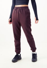 Bordeauxfarbene Jogginghosen mit seitlichen Akzenten, schmal zulaufenden Bündchen und einem elastischen Kordelzugbund. Getragen mit dunklen Schuhen und einem enganliegenden Oberteil.