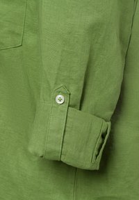 Dettaglio di una camicia di lino verde, con un bottone sul polsino, texture liscia e una tasca arrotondata sulla manica con cuciture visibili.