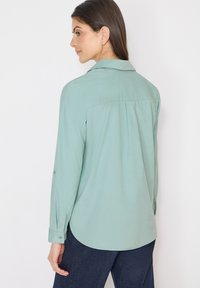 Femme aux cheveux bruns portant une chemise à manches longues vert clair et un pantalon bleu foncé, légèrement de profil sur un fond blanc.