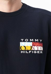 Mørkeblå sweatshirt med ribbestrikket Crew-neck. Har et flerfarget logo-merke med "TOMMY HILFIGER" og et tall-design på brystet.