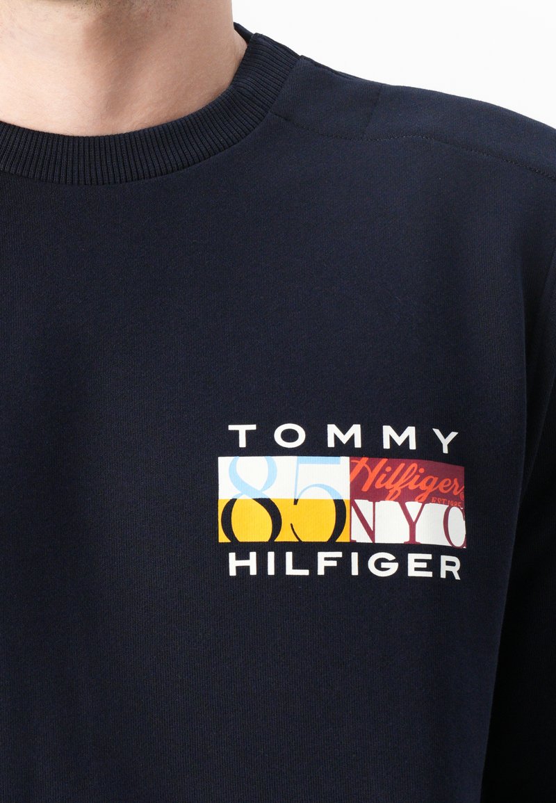 Felpa blu marino con collo a rovescio rigato. Presenta una toppa con logo multicolore con "TOMMY HILFIGER" e un design numerico sul petto.