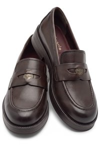 Paire de mocassins en cuir marron foncé avec un bout arrondi et un emblème décoratif en métal sur la bride, présentée sur fond blanc.