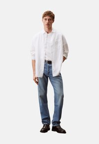 Camisa blanca de botones, camiseta blanca lisa debajo, jeans de mezclilla azul claro, cinturón negro y zapatos negros. Corte recto, tejido suave, diseño minimalista.