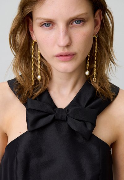 Abito in raso nero con un grande fiocco al colletto. La modella indossa lunghe orecchini a catena dorata con un accento di perla. Trucco leggero e naturale e capelli mossi.