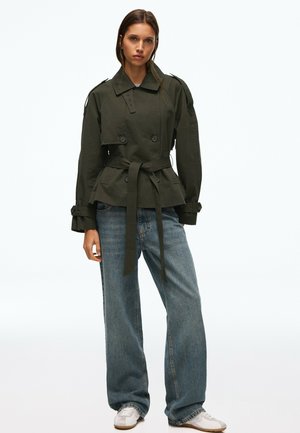 Kvinde iført en olivengrøn jakke med bælte, løse blå jeans og hvide sneakers, stående mod en ensfarvet baggrund.