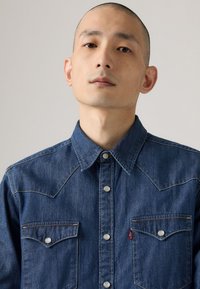 Camisa de mezclilla azul oscuro con collar puntiagudo, botones a presión y dos bolsillos en el pecho. Presenta costuras de contraste y una etiqueta de la marca en el bolsillo izquierdo.