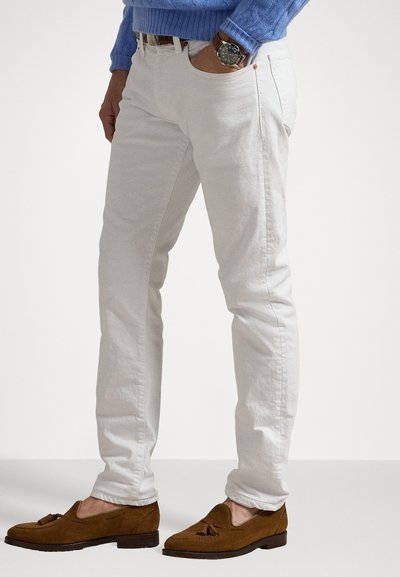 Muškarac nosi bijele uske hlače, smeđe suède mokasine s rese, plavi džemper s pletenim uzorkom i ručni sat s kožnom narukvicom.