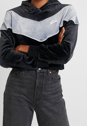 Personne portant un sweat à capuche en velours Nike noir et gris, bras croisés, et un jean taille haute gris foncé devant un fond uni.