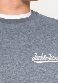Primer plano de una persona que lleva una sudadera gris de Jack & Jones encima de una camiseta blanca de cuello redondo, contra un fondo liso.