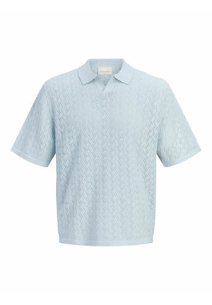 Băiat zâmbitor în picioare, purtând un pulover texturat cu mâneci scurte de culoare albastru deschis, pantaloni scurți bej, șosete albe și pantofi maro deschis cu șireturi pe fundal alb.