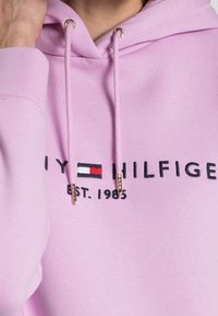 Rosa Kapuzenpullover mit Kordeln mit goldenen Metallenden, besticktem Logo und dem auffällig dargestellten Text "TOMMY HILFIGER EST. 1985".