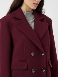 Donna che indossa un cappotto di lana bordeaux scuro doppiopetto sopra un maglione a collo alto grigio chiaro a coste.