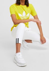 Gul bomullströja med vitt logo, vita träningsbyxor med svarta ränder vid fållen, och vita sneakers med grå detaljer.