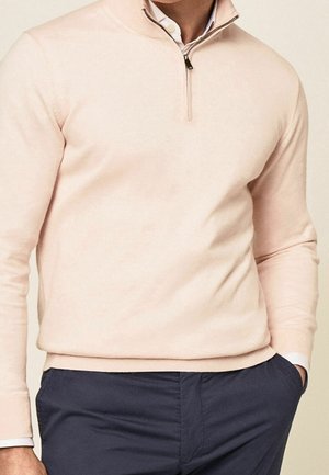 Jasnoróżowy sweter z półzamkiem przy kołnierzu, miękką fakturą i ściągaczem przy dole. Nosi się go na białą koszulę z kołnierzykiem i łączy z granatowymi spodniami.