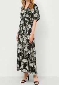 Blomstret maxi-kjole i sort, med hvide og lysegrønne blomstermønstre, korte ærmer og en afslappet pasform. Stoffet er glat.