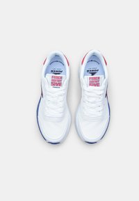 Baskets blanches avec une tige en cuir lisse, semelle bleue, accents rouges sur le talon et une étiquette bien visible indiquant "FORCE 2WENTY FIVE."