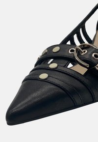 Crna cipele s oštrim vrhom od teksturirane sintetičke kože, s preklapajućim trakama, metalnim alkemijom i srebrom naglašenim prstenom.