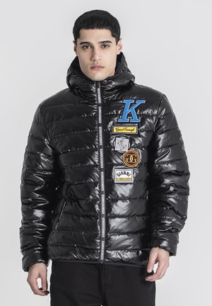 Chaqueta negra acolchada con acabado brillante, capucha y múltiples parches en azul, amarillo y marrón. Presenta cierre de cremallera y acentos de cinta de marca.