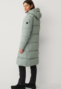 Lang, puffy grøn frakke med hætte, der har lodrette quiltede mønstre og et sort logo-patch på overarmen. Båret med mørke jeans.
