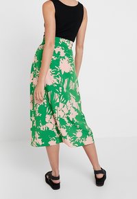 Blumiger Midi-Rock mit grünem und pinkem Muster, aus leichtem Stoff gefertigt. Kombiniert mit einem schwarzen gerippten Tanktop und schwarzen Sandalen.