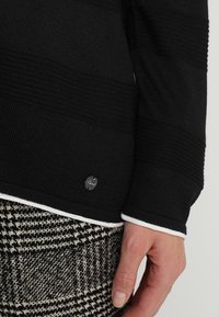 Czarny sweter z fakturowanymi paskami, z białym wykończeniem przy dekolcie i mankietach. Mała metka z logo jest widoczna na dole. Czarno-białe w kratę spodnie dopełniają całość.