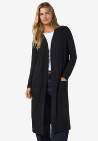 Svart lang cardigan med lange ermer, åpen front, to lommer foran og ribbet tekstur; kombinert med en hvit topp og denimjeans.