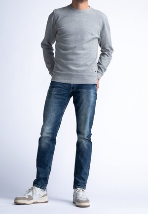 Mann trägt hellgrauen Sweatshirt, blaue Jeans und weiße Sneaker, steht mit den Händen hinter dem Rücken vor weißem Hintergrund.