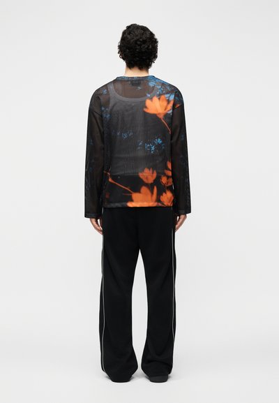 Homme aux cheveux bouclés, vu de dos, portant une chemise noire en maille à manches longues avec un imprimé floral orange et un pantalon noir à rayures blanches.
