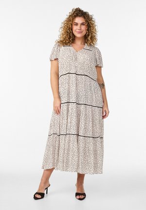 Kvinde med krøllet hår iført en beige, kortærmet midi-kjole med sorte prikker og sorte åbne sandaler, stående mod hvid baggrund.