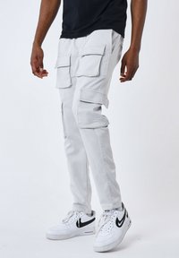 Pantalones cargo en tela gris claro con múltiples bolsillos con solapa, ajuste ajustado y cremalleras; combinados con zapatillas blancas con detalles en negro.