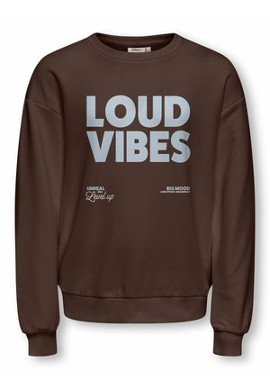 Fille aux cheveux bouclés portant un sweat-shirt marron avec le texte "LOUD VIBES", un jean gris large et des baskets blanches, debout avec la main sur la hanche.