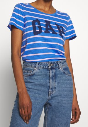 Person iført en blå og pink stribet t-shirt med teksten "GAP", kombineret med højtaljede blå denimjeans mod en ensfarvet baggrund.
