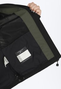 Black jacket interior with green accents, mesh lining, and a label featuring name and contact fields. Adjustable drawstring at the bottom.