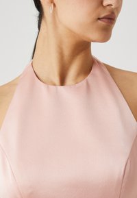 Halter-neck dress in light pink fabric with a smooth texture; features a rounded neckline and subtle sheen. Includes small hoop earrings.