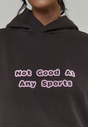 Gros plan sur un sweat à capuche sombre avec un texte brodé rose "Pas bon dans aucun sport" sur la poitrine.
