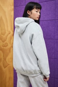 Una giovane donna con capelli neri che indossa un hoodie grigio chiaro e pantaloni della tuta è in posizione laterale contro un muro viola e di legno.