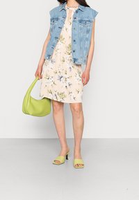 Džínsová bezrukávová bunda přes květinové světle béžové šaty. Nese zelenou kabelku a má na sobě odpovídající zelené sandály.