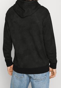 Black hooded sweatshirt with a soft, textured finish. Features ribbed cuffs and hem, and a relaxed fit. Worn with light blue denim jeans.