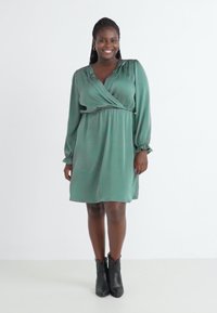 Vihreä kietaisuhelma pitkähihaisilla, vyötärölle kiristetty ja sileä rakenne, yhdistettynä mustiin nilkkasaappaisiin. Yksinkertainen, polvenpituinen muotoilu.