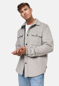 Giacca in pile grigia con design con colletto, chiusura frontale con bottoni, due tasche sul petto e orlo dritto. Indossata sopra una t-shirt bianca.