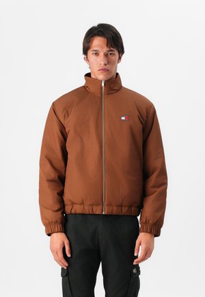 Chaqueta acolchada marrón con cuello alto, cremallera frontal, puños elásticos y un pequeño logo bordado en el lado izquierdo del pecho. Pantalones negros puestos.