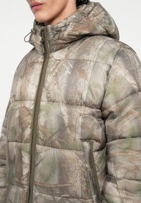 Kamouflage steppelt dzseki barna és zöld árnyalatokban. Kapucnival, cipzáros záródással és fás textúrájú anyaggal rendelkezik.