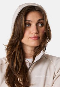 Light beige hoodie with a high collar and partial zipper, featuring a white brand logo on the chest and a soft, textured fabric.