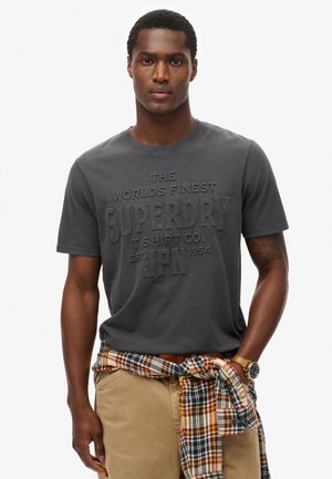 Gray cotton t-shirt featuring embossed text, "Superdry," with a round neckline. Paired with khaki pants and a checkered flannel shirt tied at the waist.