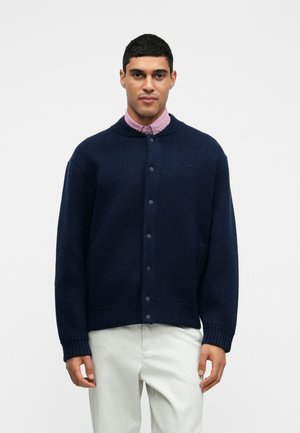 Navy blue knit cardigan with a ribbed collar, button closure, and side pockets. Worn over a light pink shirt, featuring a subtle logo detail.